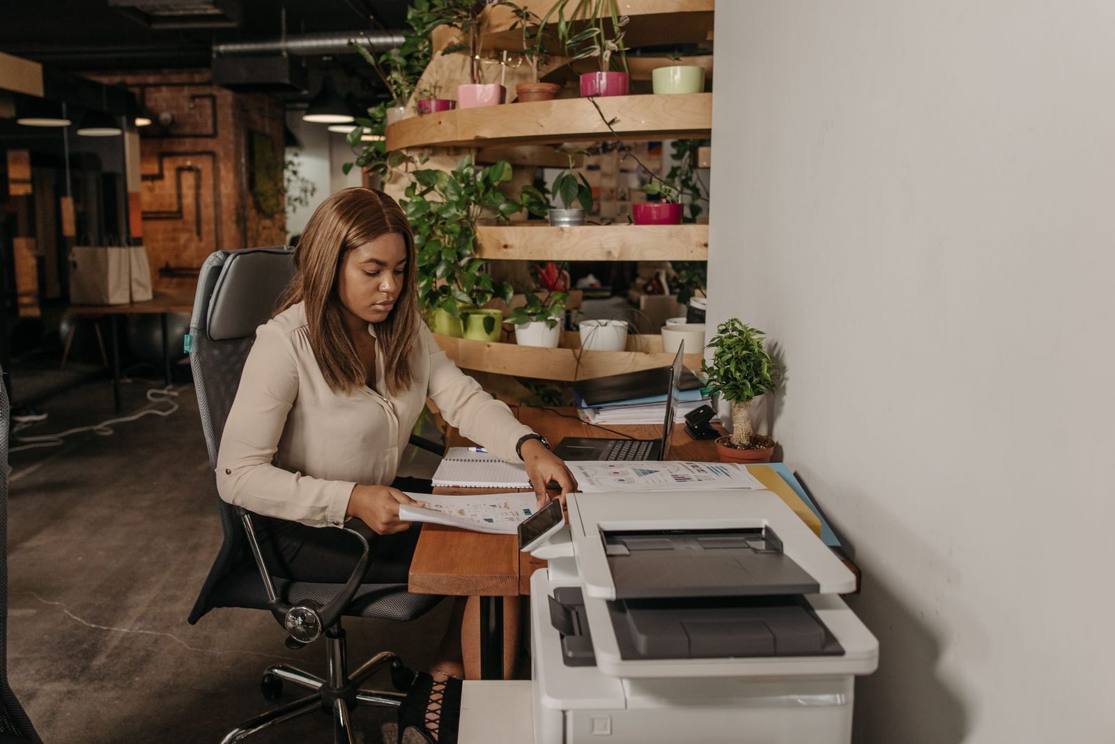 Mujer sentada a un escritorio revisando documentos en papel junto a una impresora multifunción y un ordenador portátil