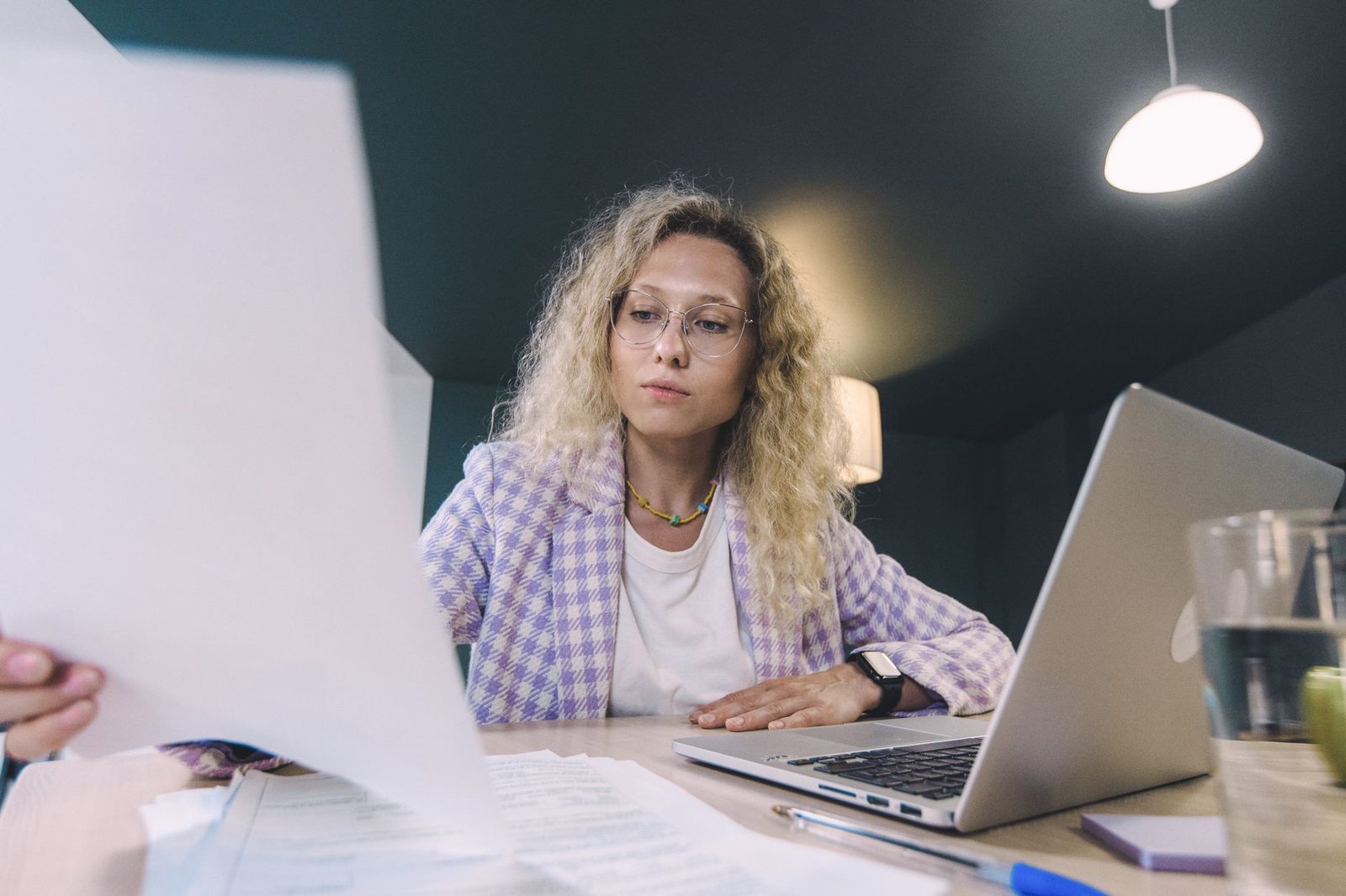 Woman wearing glasses in front of a laptop, reviewing paper documents on a desk