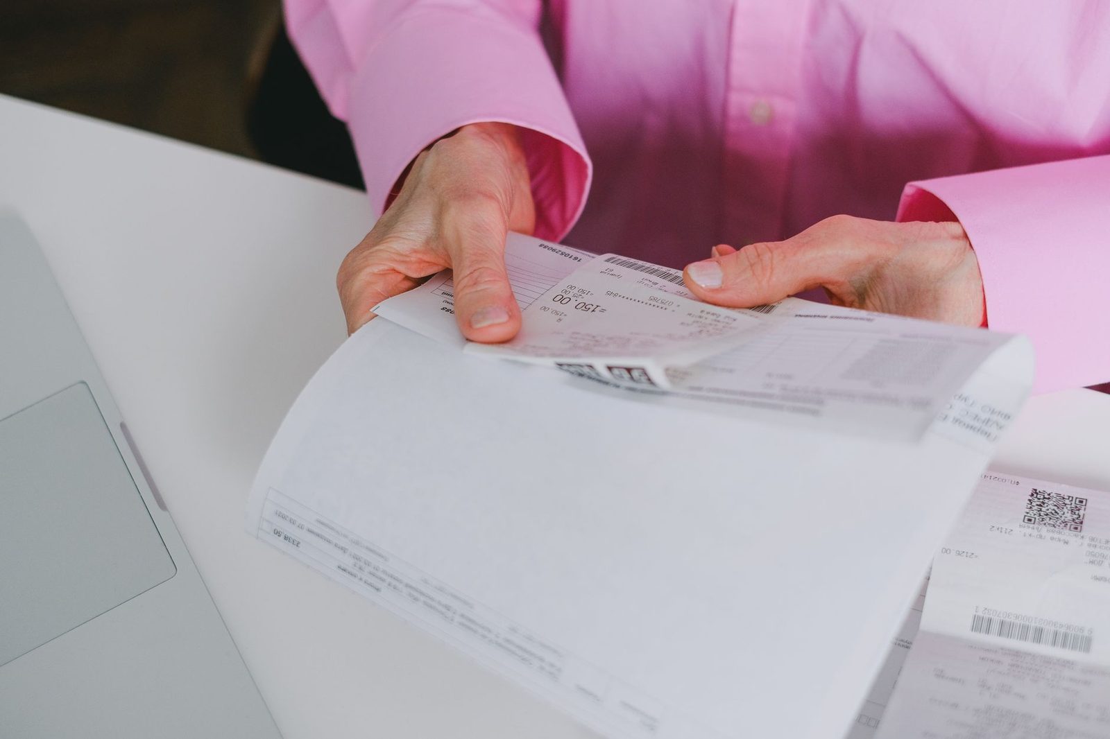 Mains tenant des factures papier avec codes-barres sur un bureau blanc, à côté d'un ordinateur portable