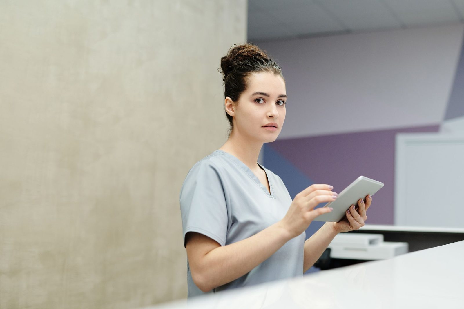 Soignante en tenue bleue utilisant une tablette numérique derrière un comptoir d'accueil médical