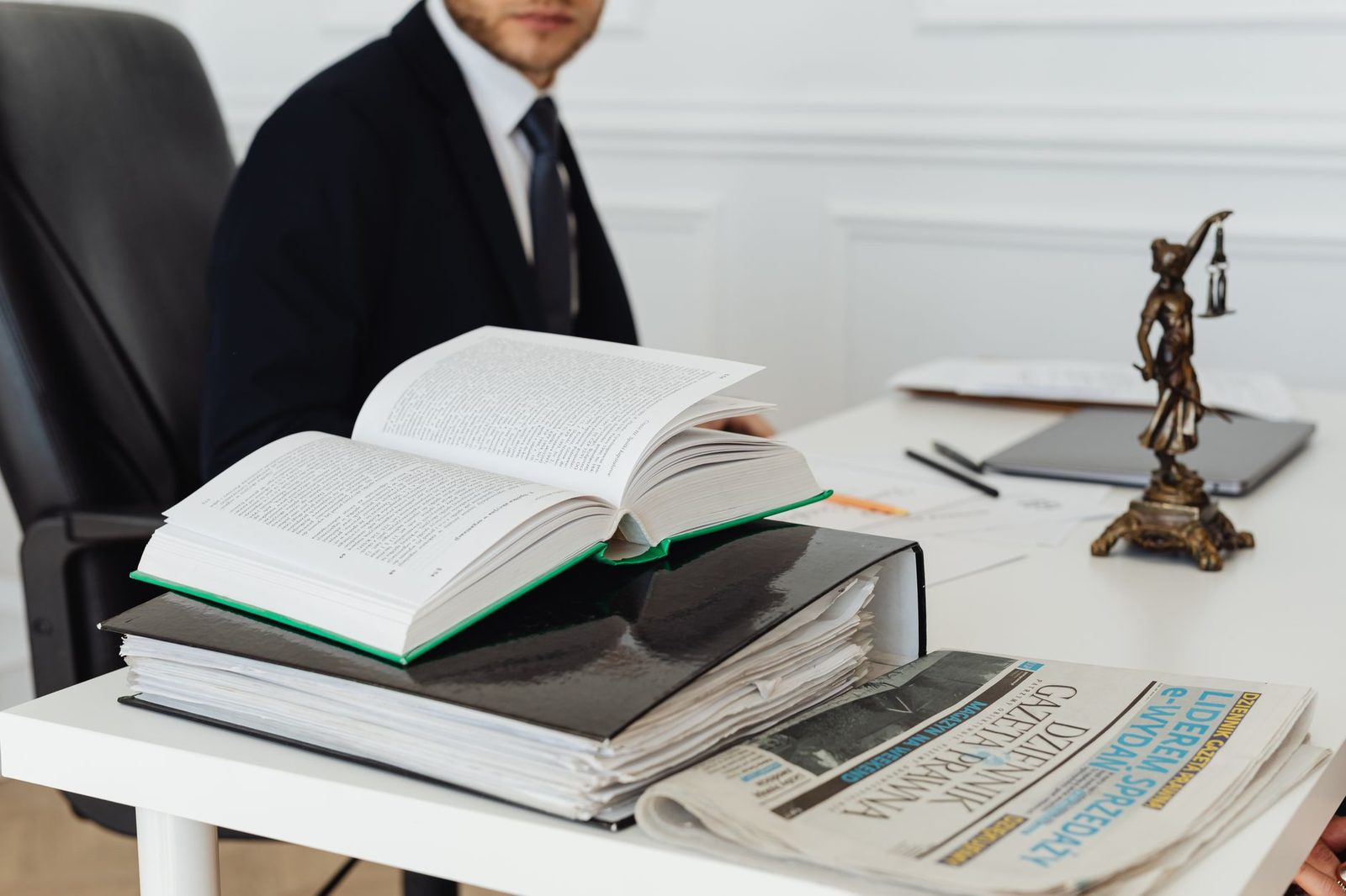 Livre de droit ouvert sur un bureau avec journaux, statue de la justice et homme en costume assis