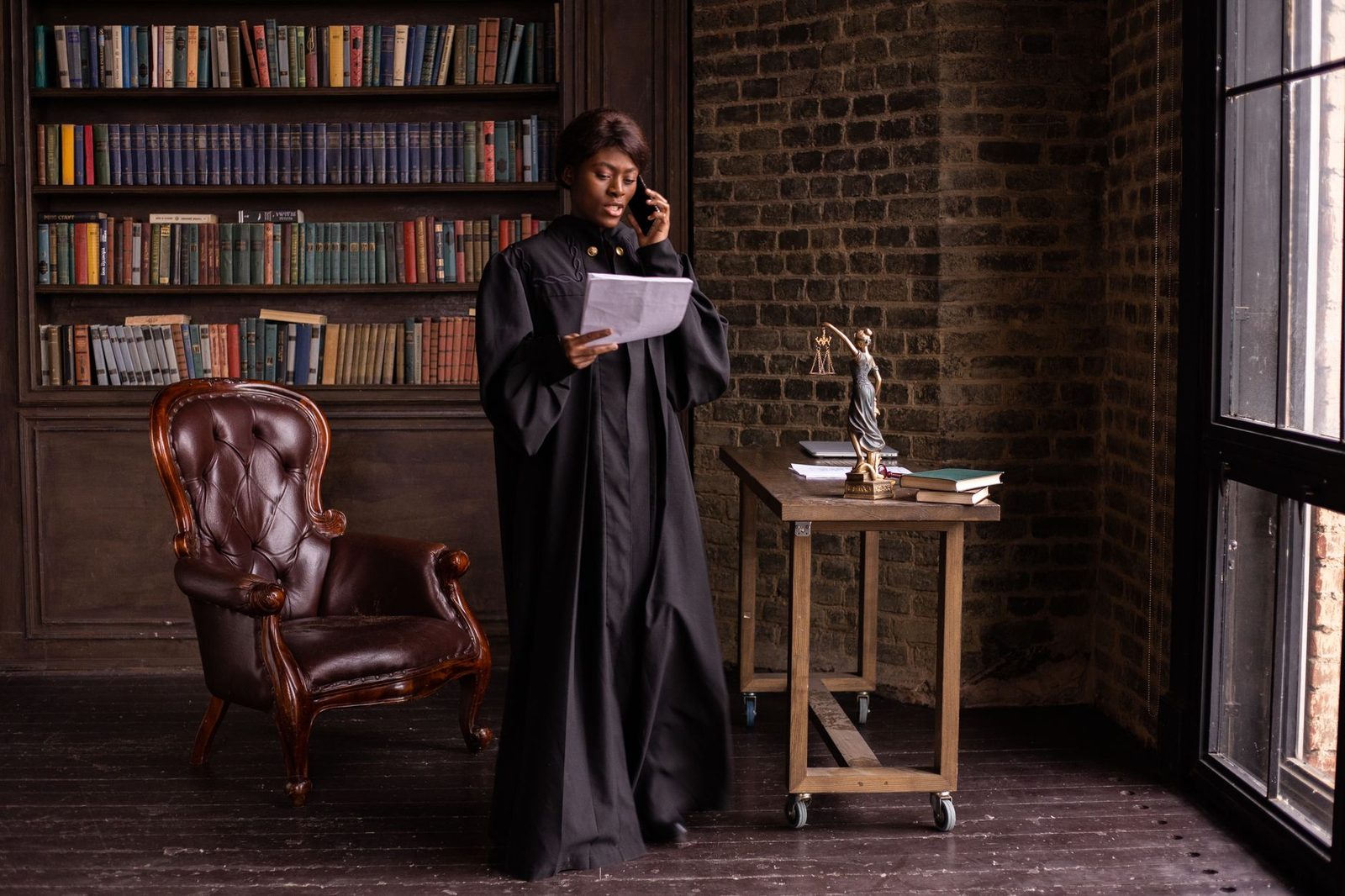 Femme en robe d'avocate au téléphone lisant des documents devant une bibliothèque et une statuette de la justice