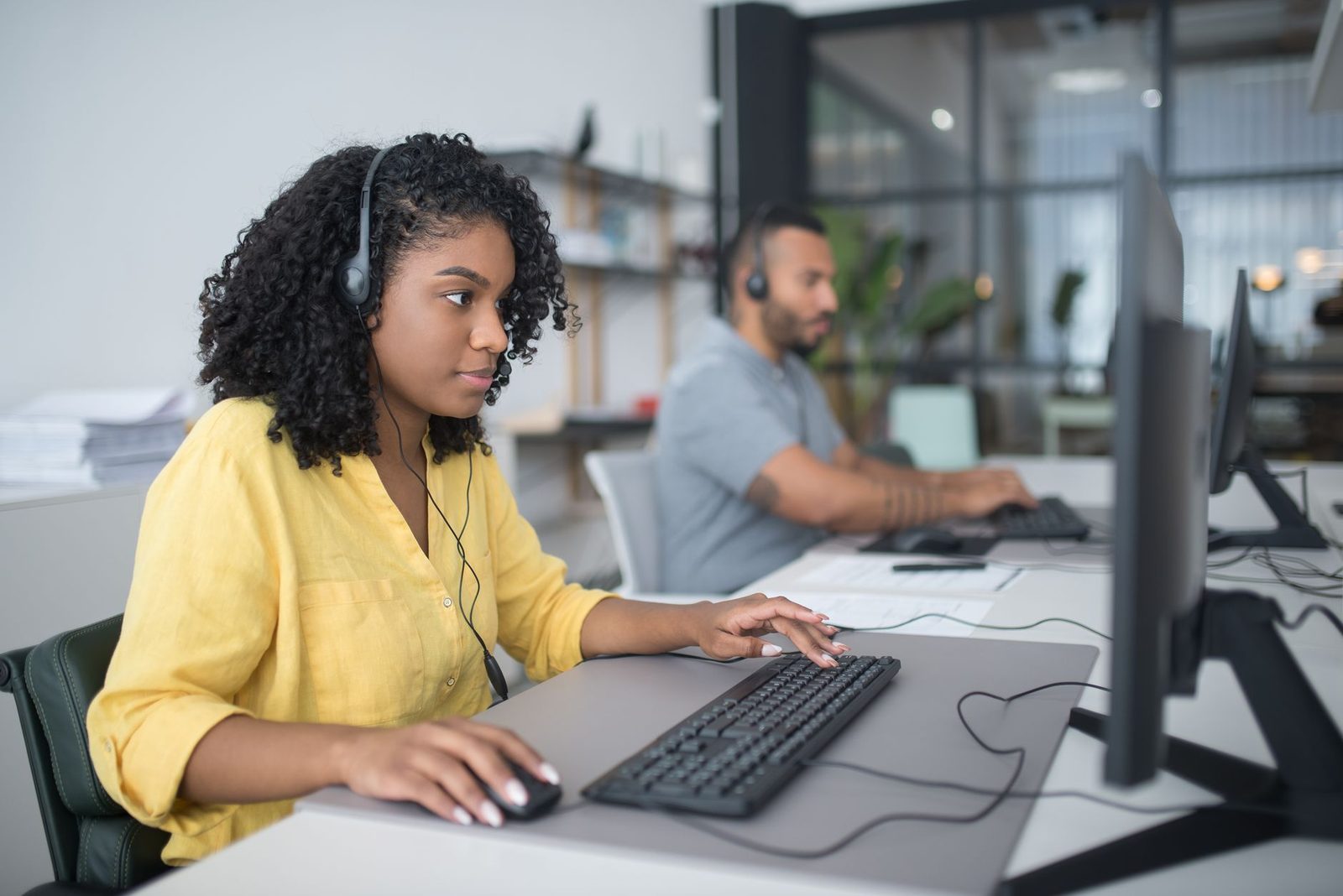 Femme avec casque téléphonique devant un ordinateur dans un bureau, un collègue au second plan