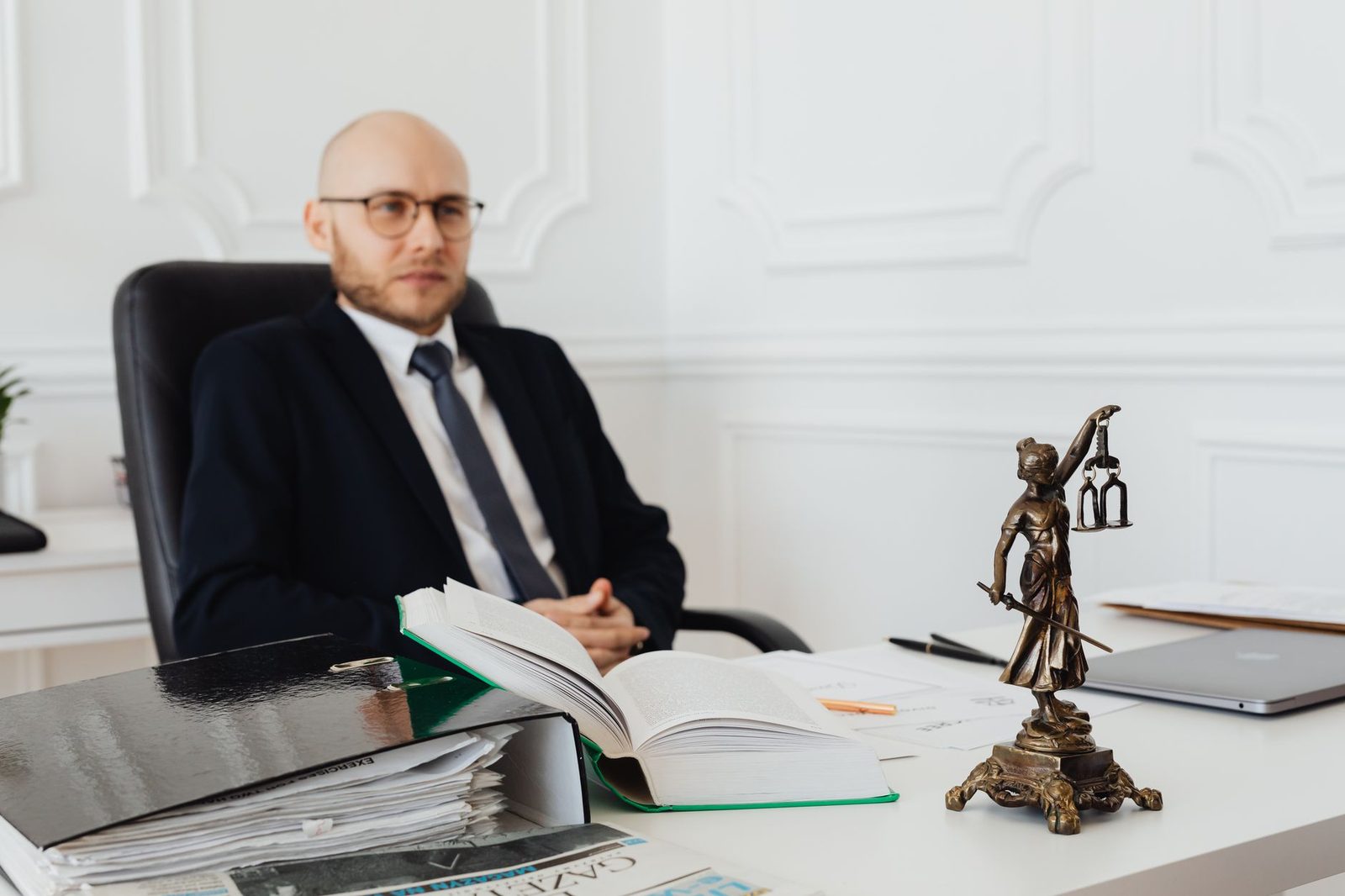 Juriste en costume assis à son bureau avec un livre de droit ouvert, une statuette de la justice et un ordinateur portable