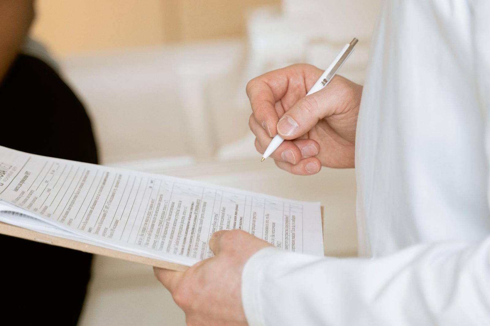 Healthcare professional in a white coat filling in a paper form on a clipboard with a pen