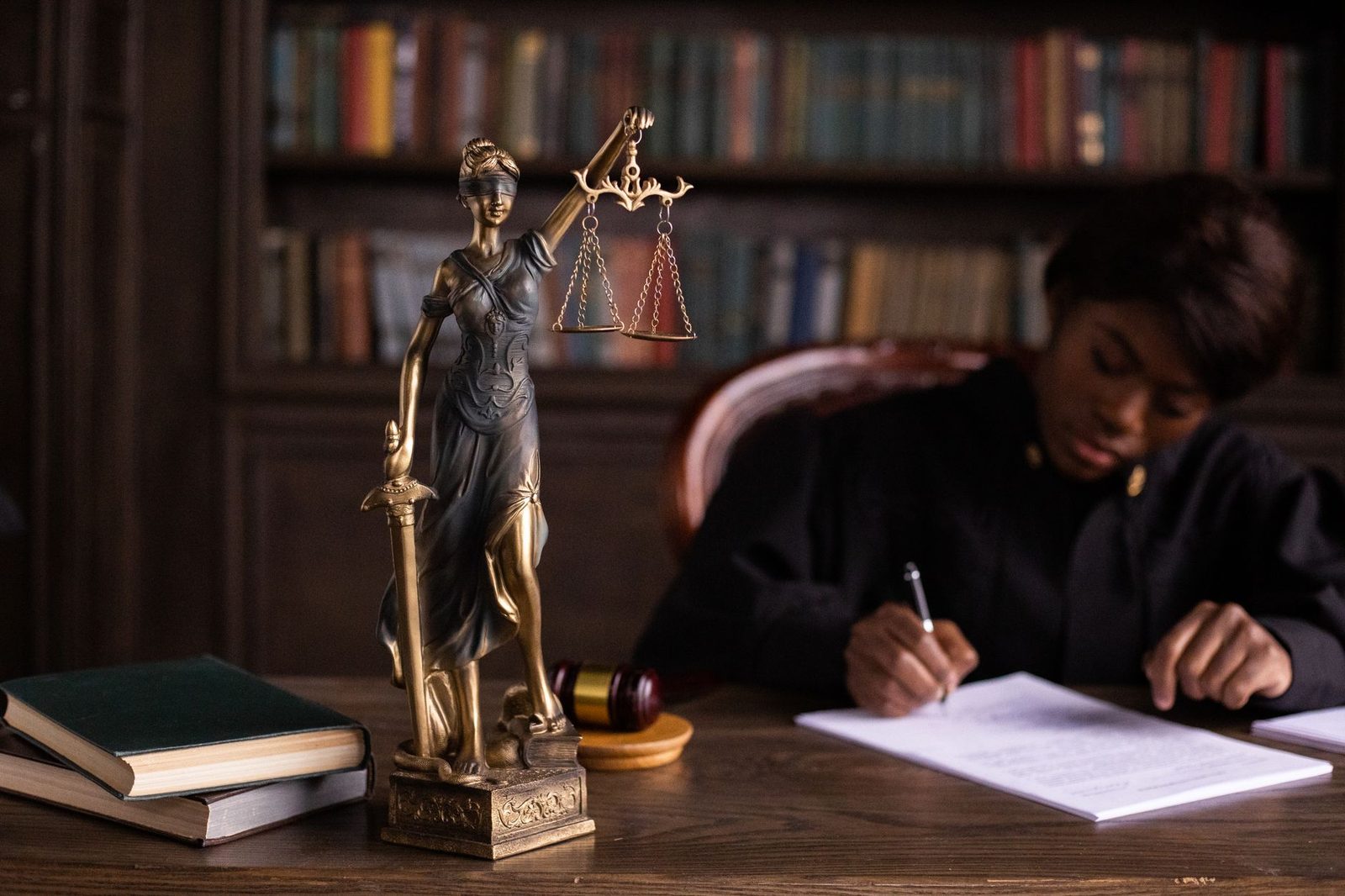 Statue de la justice et maillet sur un bureau, femme en robe noire écrivant devant une bibliothèque juridique