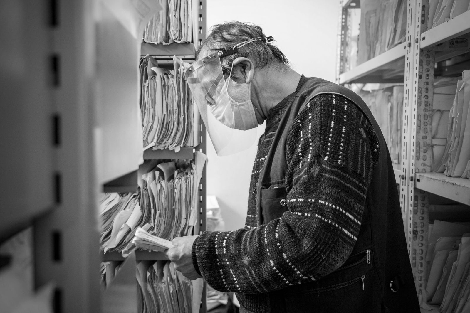 Hombre con mascarilla consultando expedientes en papel en las estanterías de un archivo, fotografía en blanco y negro