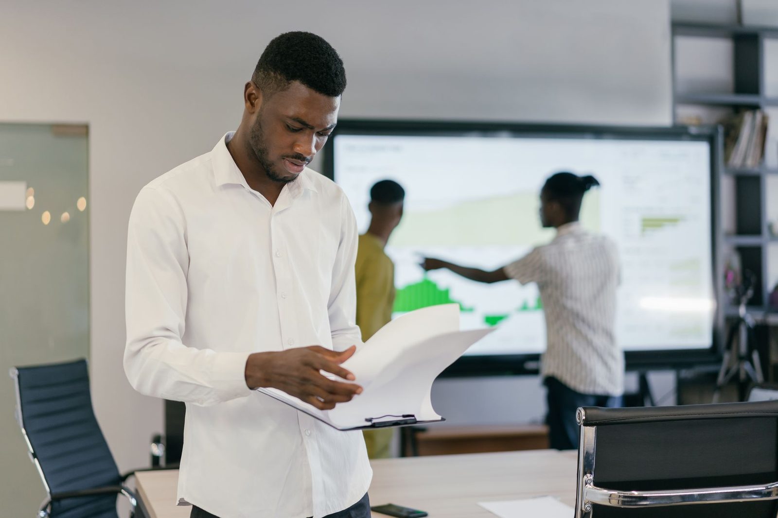 Man in a white shirt looking over paper documents in an office, colleagues in front of a screen