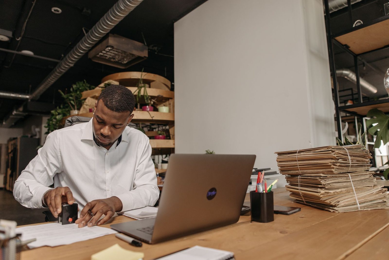 Man in a white shirt stamping a paper document at a desk, in front of a laptop and stacks of files